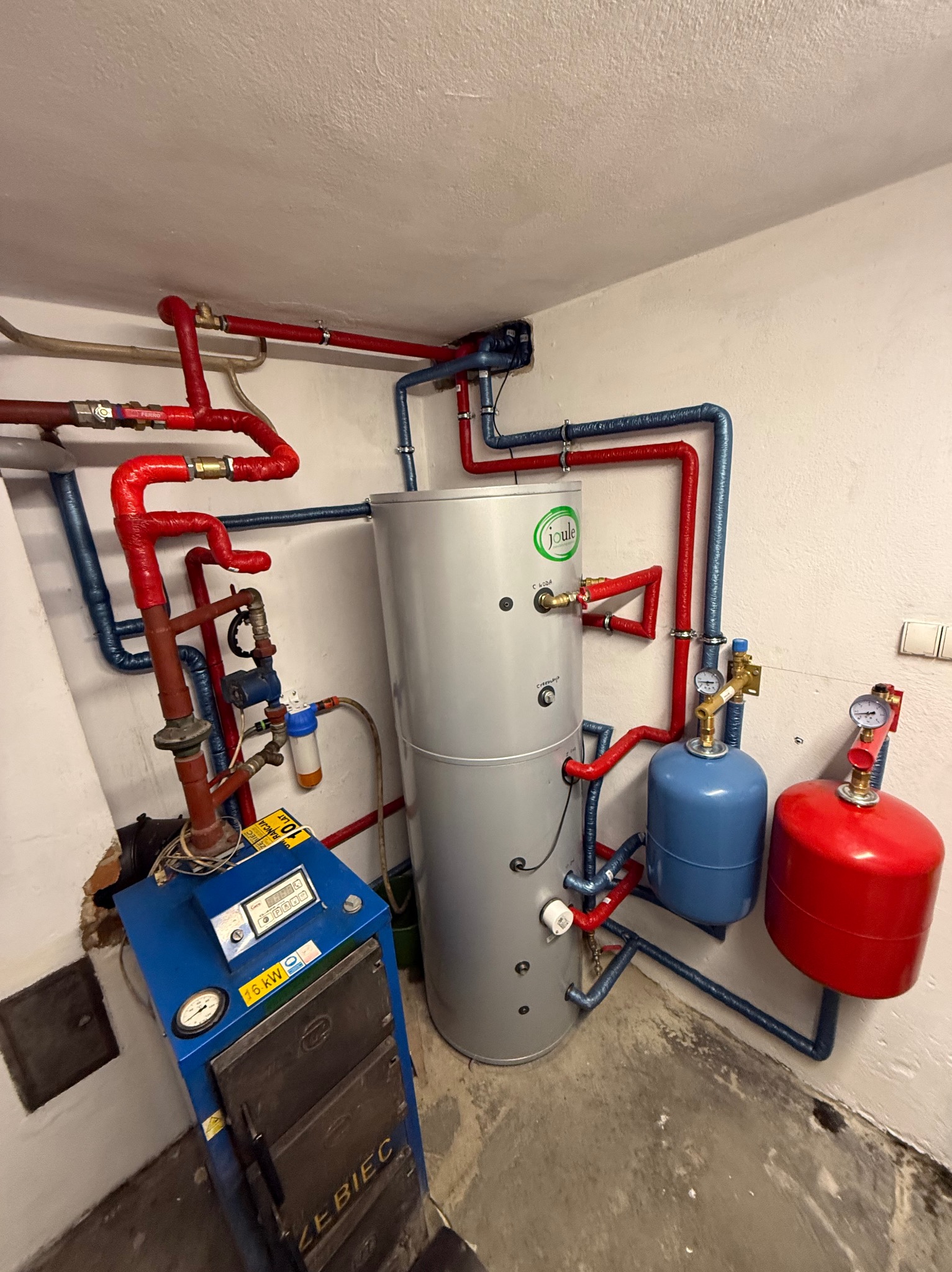 Kotłownia z piecem Zębiec 16kW, zbiornikiem Joule i instalacją z rur w kolorach czerwonym i niebieskim. Widoczne manometry i filtry. Kotłownia w piwnicy.