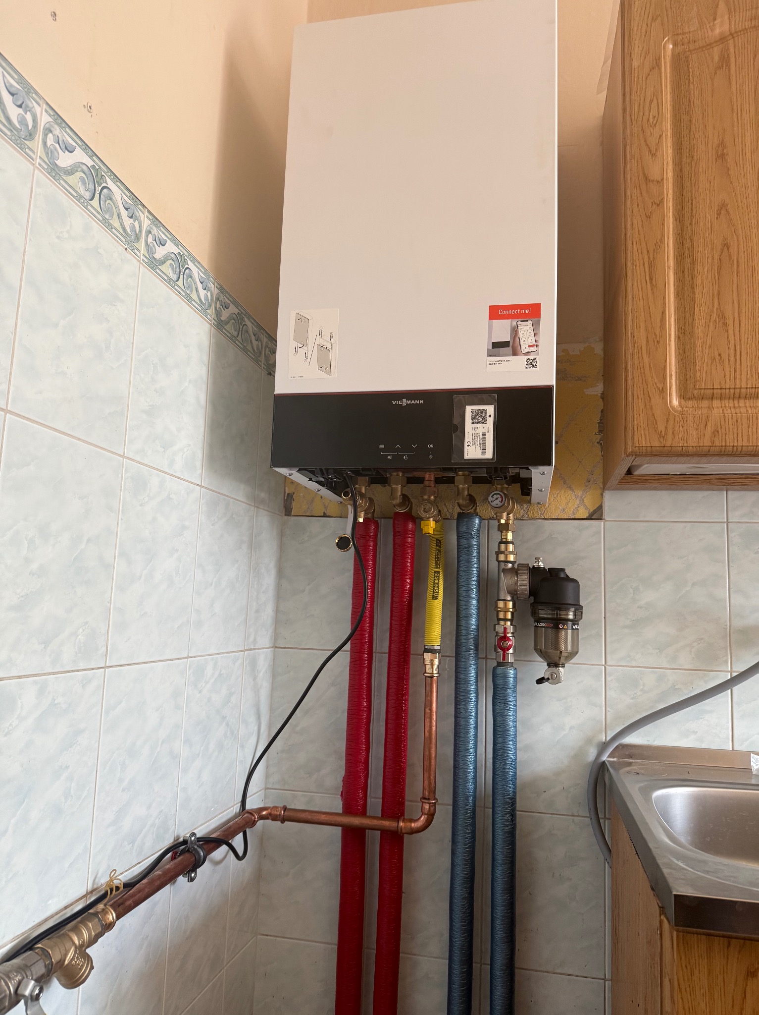 Biały piec gazowy Viessmann zamontowany na ścianie z niebieskimi płytkami, widoczne rury w kolorach czerwonym, żółtym i niebieskim, oraz filtr wody.