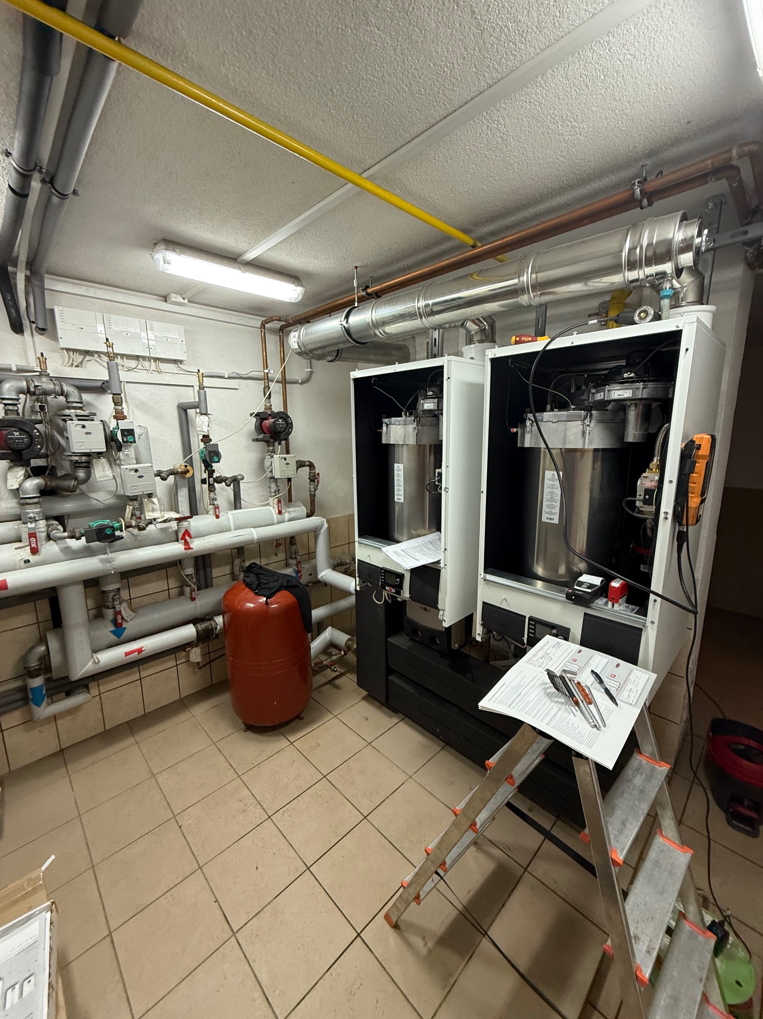 Dwa nowoczesne piece gazowe, z widoczną instalacją hydrauliczną i rurami. Na pierwszym planie drabina z dokumentami i narzędziami. Całość w pomieszczeniu z beżowymi płytkami.
