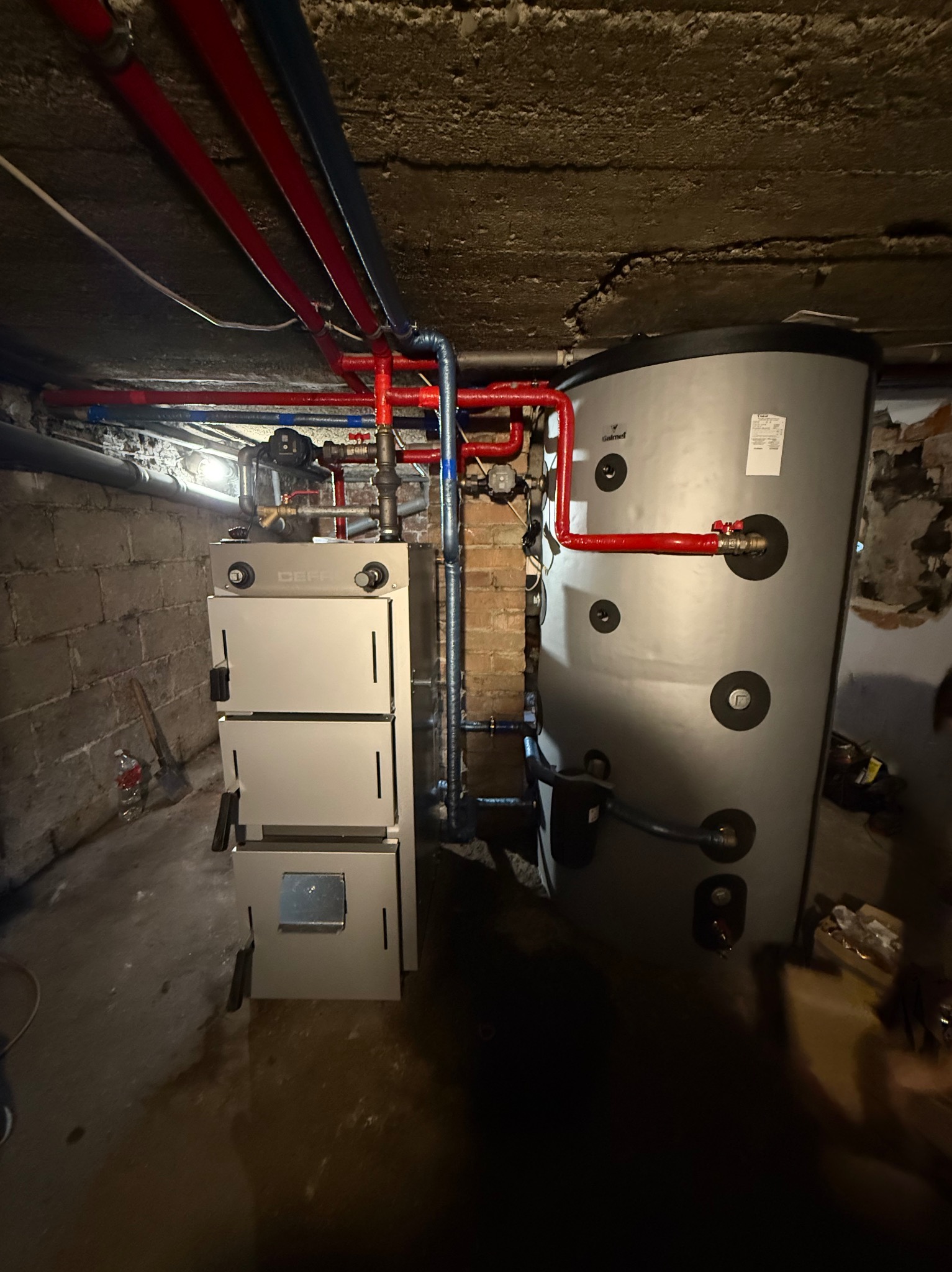 Nowoczesny piec CO z zasobnikiem w piwnicy, instalacja z rur miedzianych i plastikowych, widok na ceglany mur i betonowy sufit. CEFF, Galmet.