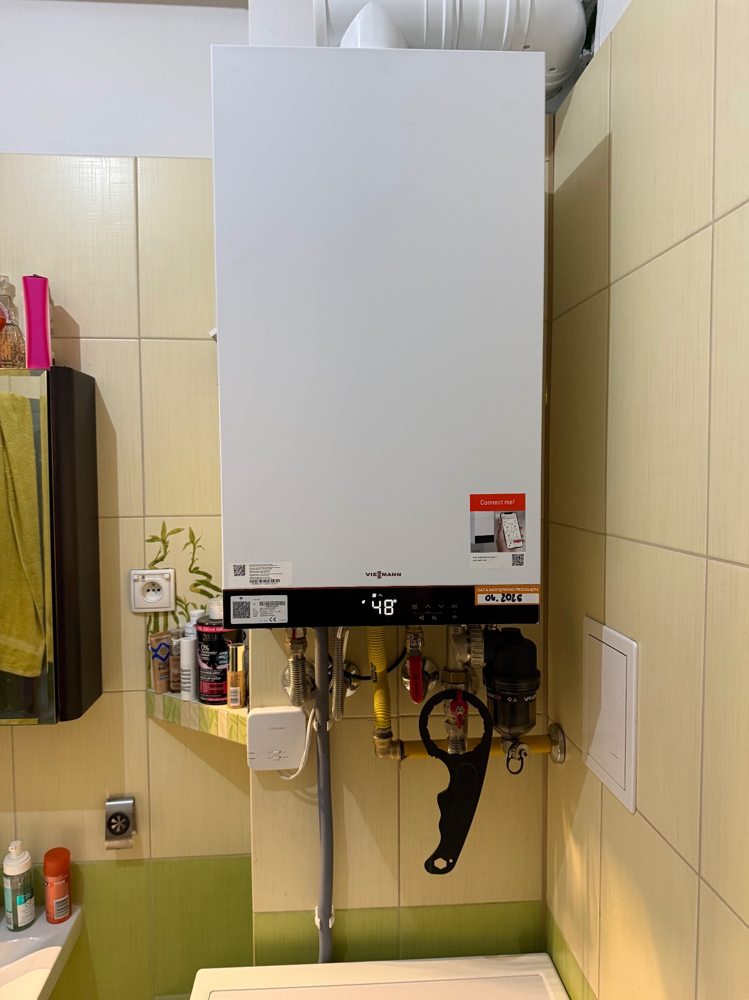 Biały piec gazowy Viessmann na ścianie z beżowych płytek, z widocznymi rurami, zaworami i narzędziem do instalacji. Wyświetlacz pieca pokazuje temperaturę 48 stopni.