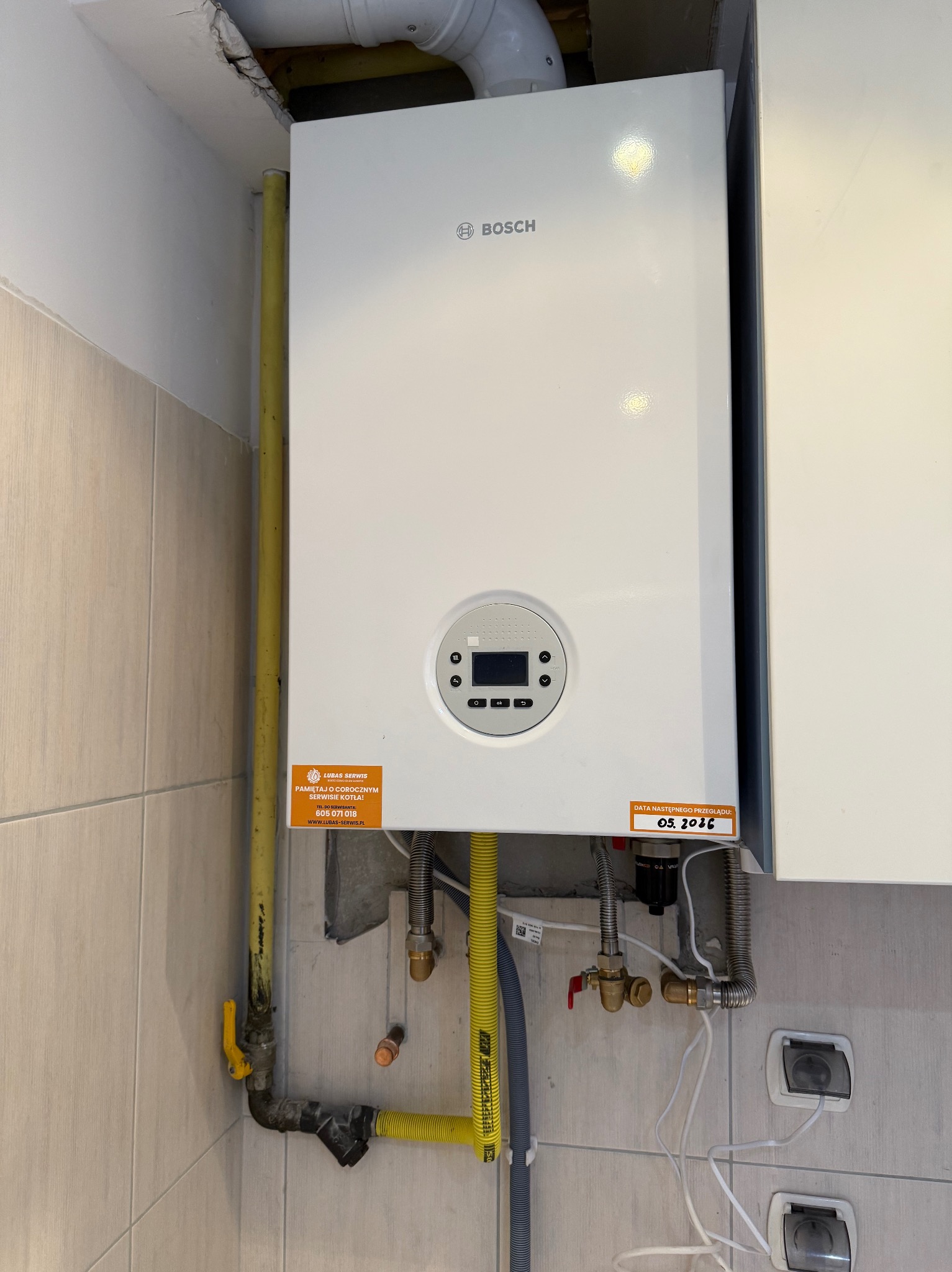Biały, wiszący piec gazowy Bosch na tle płytek. Widoczne żółte rury gazowe, naklejka informująca o serwisie i dacie przeglądu. Instalacja w pomieszczeniu.