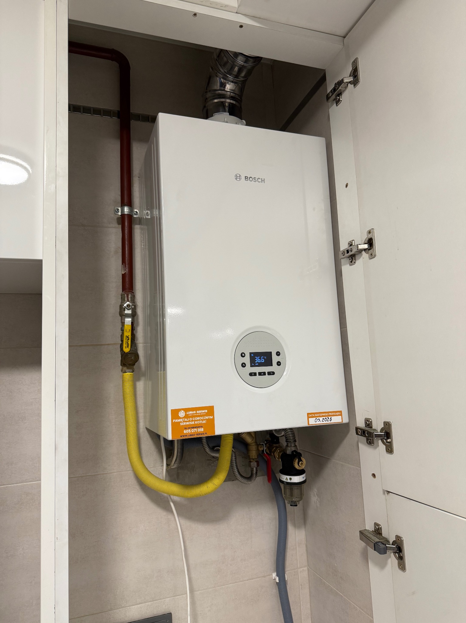 Zamontowany piec gazowy Bosch w szafce w Gwoźnicy Górnej. Widoczne przyłącza, rury, żółty wąż gazowy i naklejka z datą przeglądu.