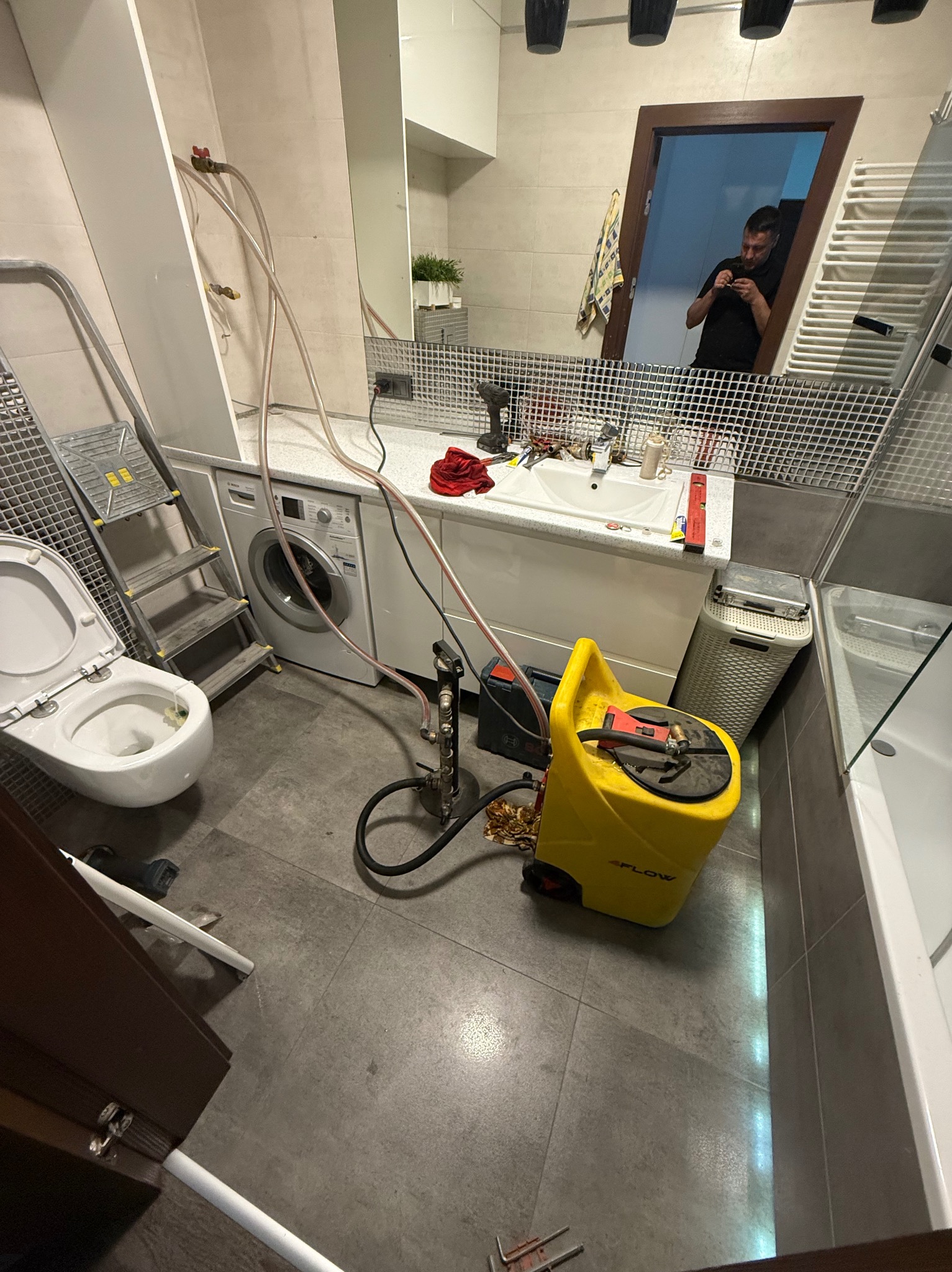 Łazienka w trakcie prac hydraulicznych: podłączona toaleta, widoczna pralka, drabina, żółty sprzęt do testowania szczelności instalacji. W lustrze odbija się sylwetka osoby.