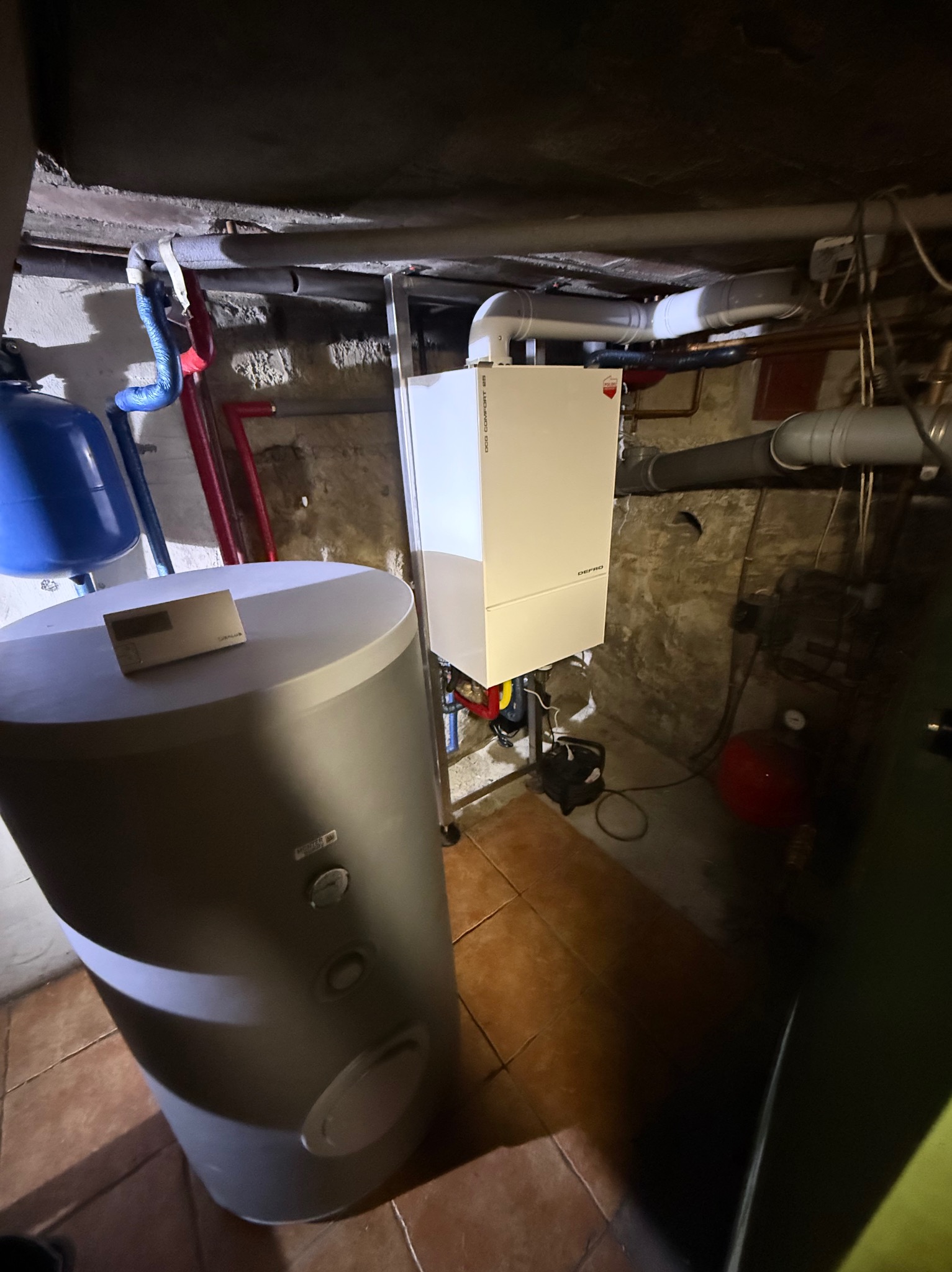 Biały piec gazowy Defro Comfort 25 w piwnicy z kamiennymi ścianami, obok szary zasobnik ciepłej wody i niebieski zbiornik wyrównawczy.