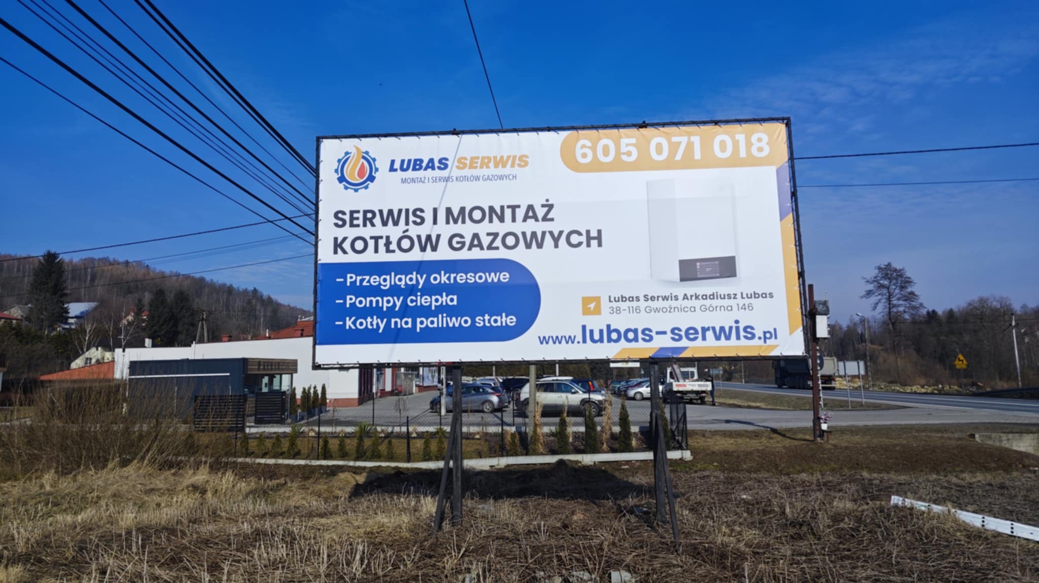 Billboard reklamujący serwis i montaż kotłów gazowych firmy Lubas Serwis, z numerem telefonu i adresem strony internetowej, na tle krajobrazu.