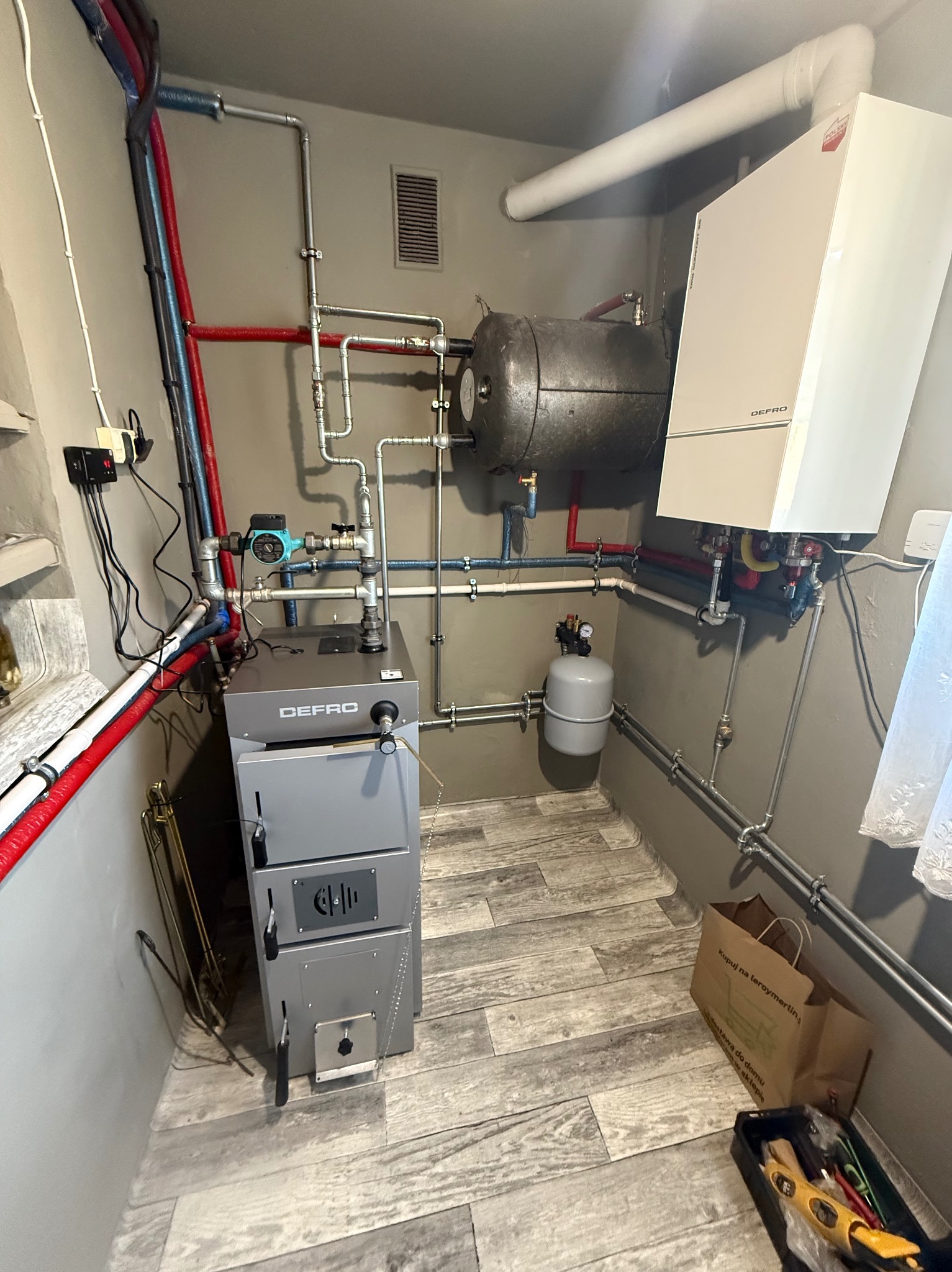 Zmontowana instalacja grzewcza z piecem Defro, zbiornikiem akumulacyjnym i osprzętem. Widoczne rury, zawory i elementy sterujące. Podłoga wyłożona panelami imitującymi drewno.
