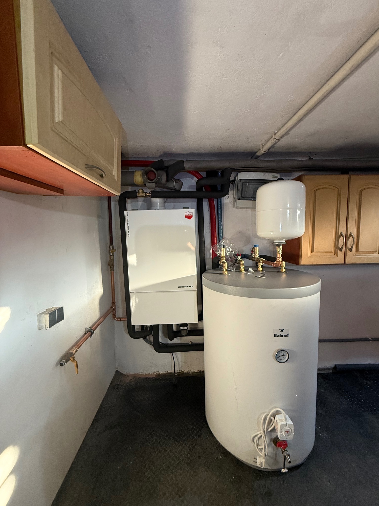 Biały piec gazowy Defrro i zasobnik Galmet w kotłowni, widoczne rury instalacji, szafki. Realizacja instalacji grzewczej, oświetlenie sufitowe.