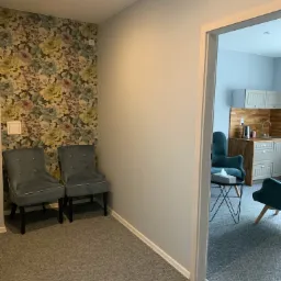 Poczekalnia: Dwa szare krzesła na tle ściany w kwiaty, widok na gabinet z aneksem kuchennym i turkusowymi fotelami