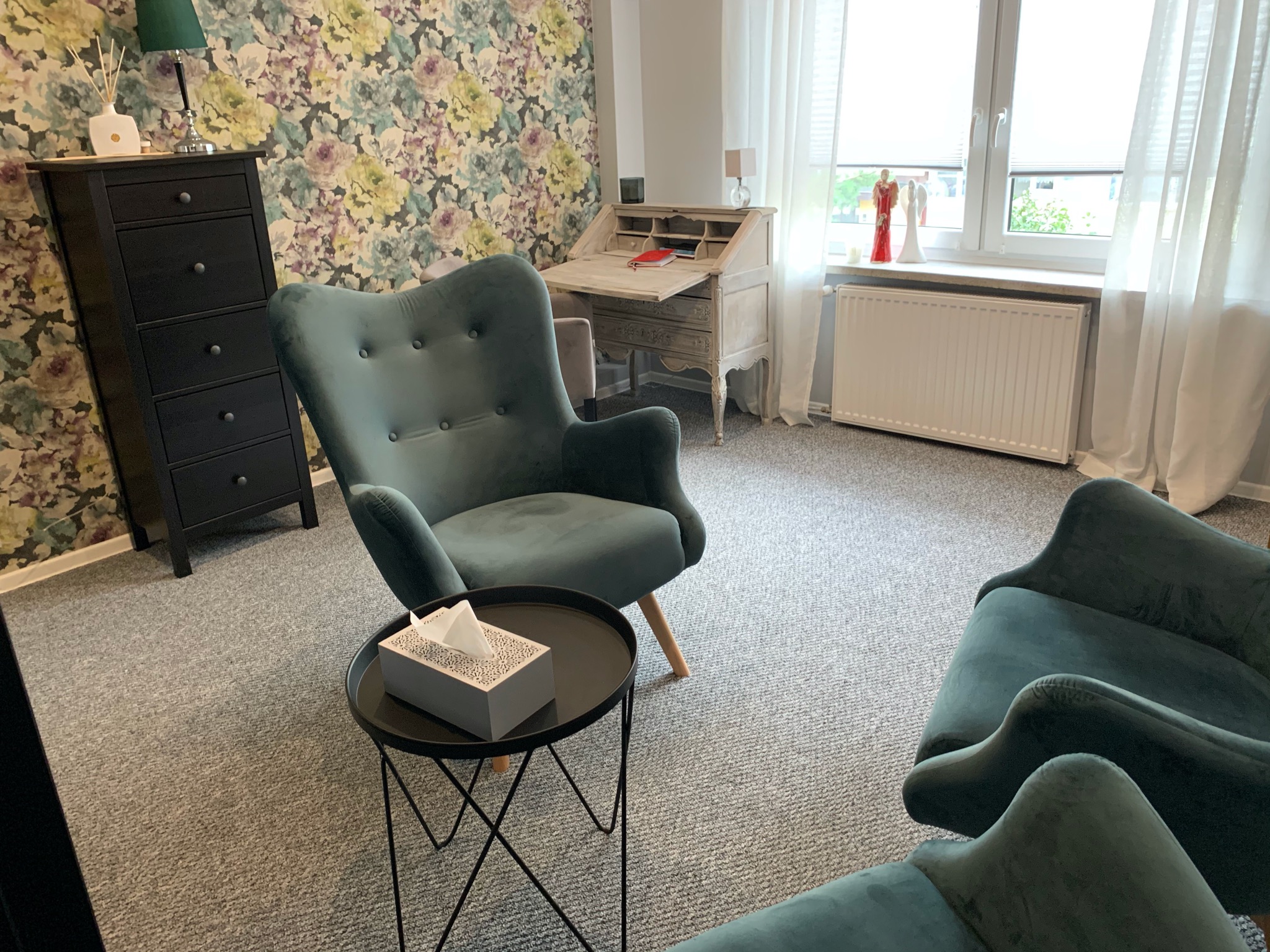 Umeblowany gabinet z dwoma zielonymi fotelami, biurkiem sekretarzykiem i komodą na tle ściany w kwiaty. Na stoliku pudełko chusteczek.