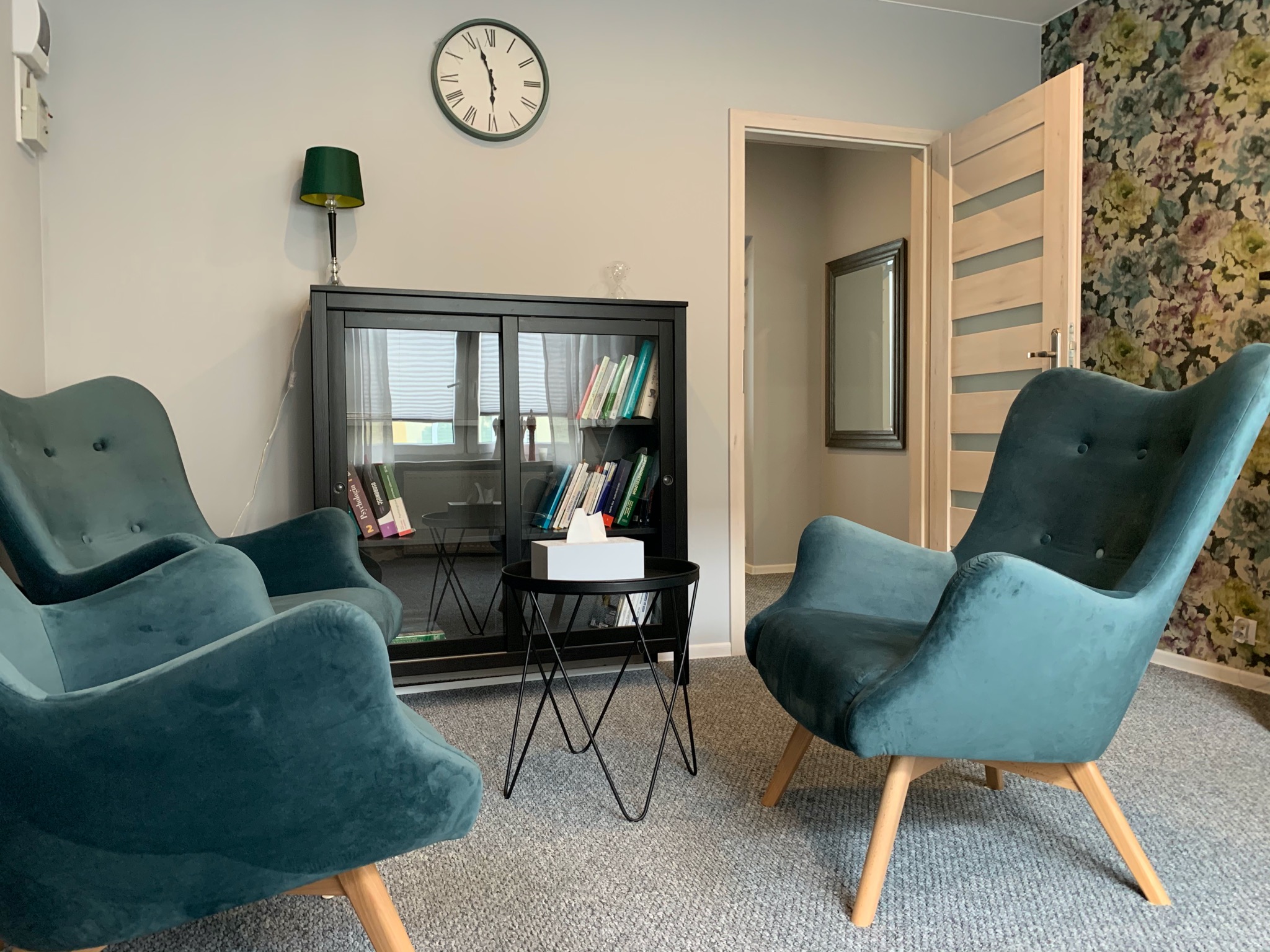 Pokój z trzema turkusowymi fotelami klubowymi, czarnym regałem z książkami, zegarem na ścianie i stolikiem kawowym z pudełkiem chusteczek.