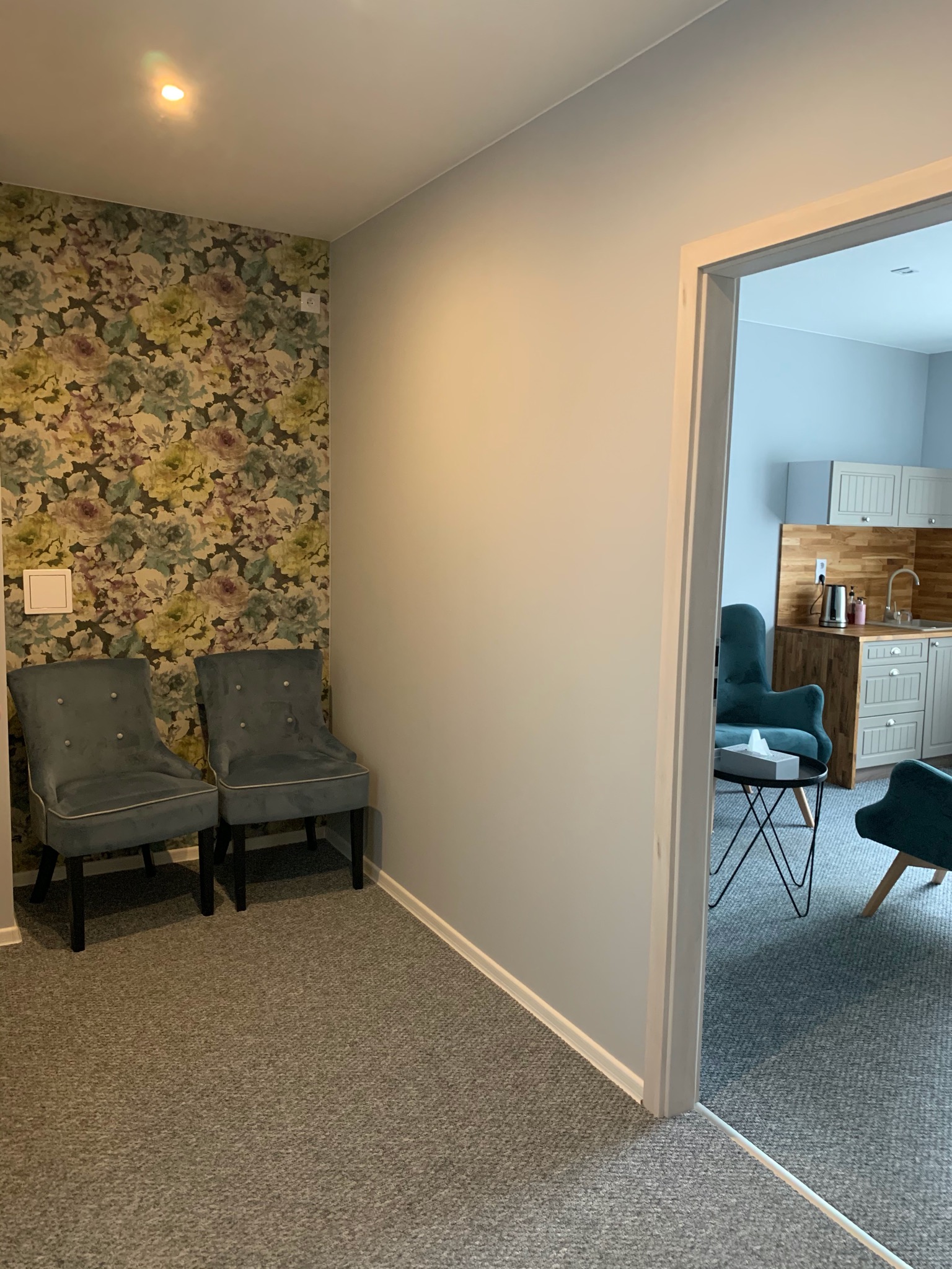 Poczekalnia: Dwa szare krzesła na tle ściany w kwiaty, widok na gabinet z aneksem kuchennym i turkusowymi fotelami