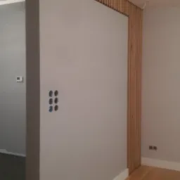 Nowoczesna aranżacja wnętrza z drewnianymi lamelami na ścianie, tworzącymi ażurową dekorację, widoczne gniazdka elektryczne i fragment drewnianej podłogi.