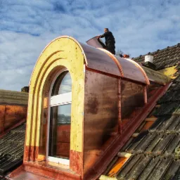 Montaż miedzianej obudowy okna dachowego typu lukarna z drewnianą konstrukcją, widoczne stare dachówki i pracownik na dachu.