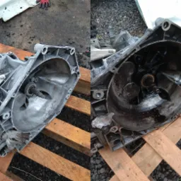 Dwie połówki aluminiowej obudowy skrzyni biegów, jedna czysta, druga brudna od oleju, leżą na drewnianej palecie na zewnątrz.