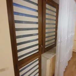 Drewniane okno z roletami w biało-szare pasy, obok okna zwiewna, biała firanka z koronkowym wykończeniem.