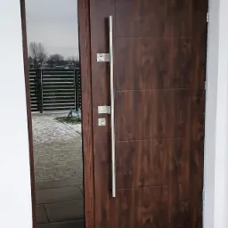Brązowe drzwi zewnętrzne z długim, srebrnym uchwytem i naświetlem bocznym, widok na posesję z kostką brukową i ogrodzeniem.