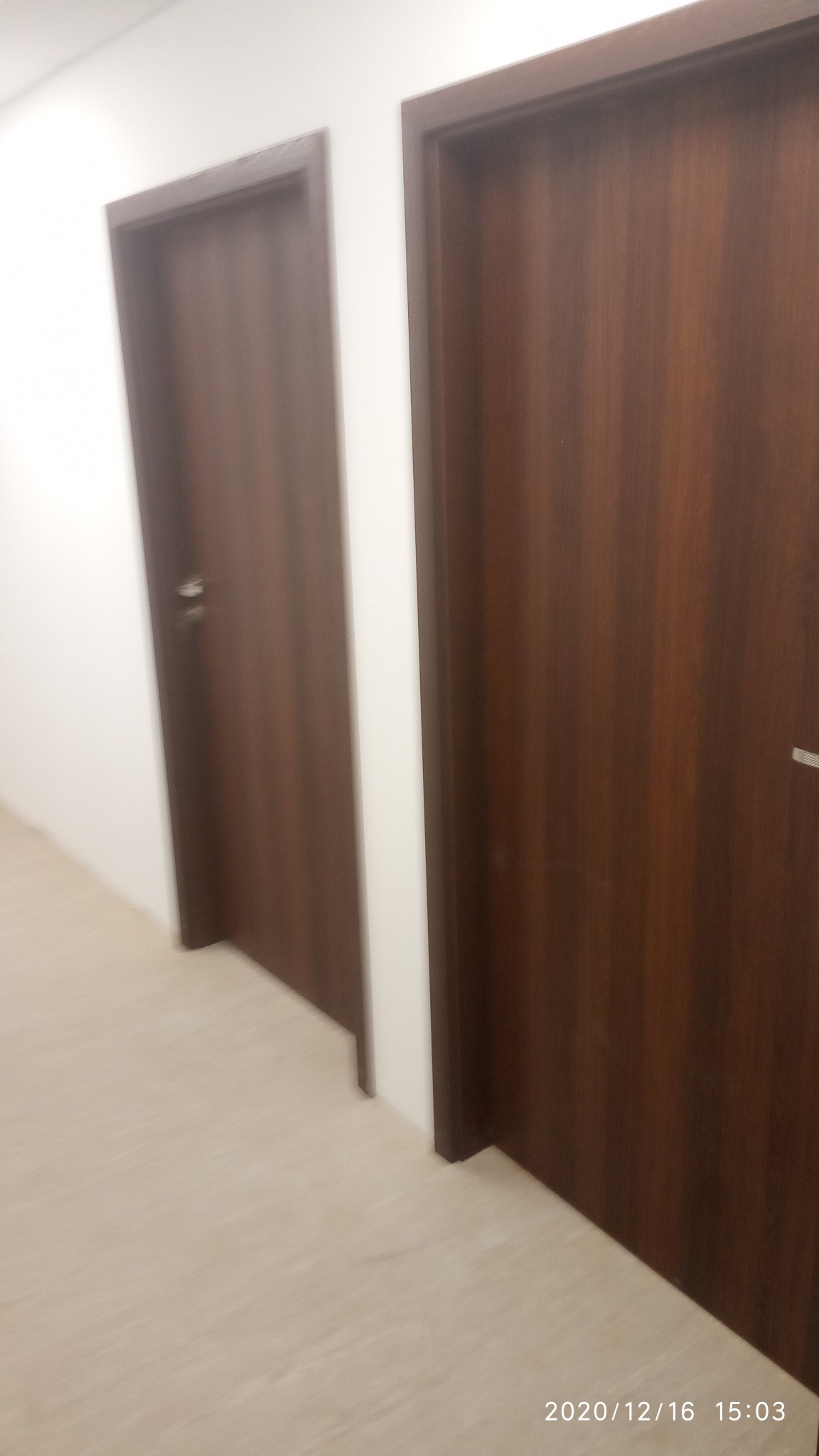 Dwa ciemnobrązowe, drewniane drzwi wewnętrzne z srebrnymi klamkami, zamontowane w białej ścianie, widoczne z perspektywy korytarza z jasną podłogą.