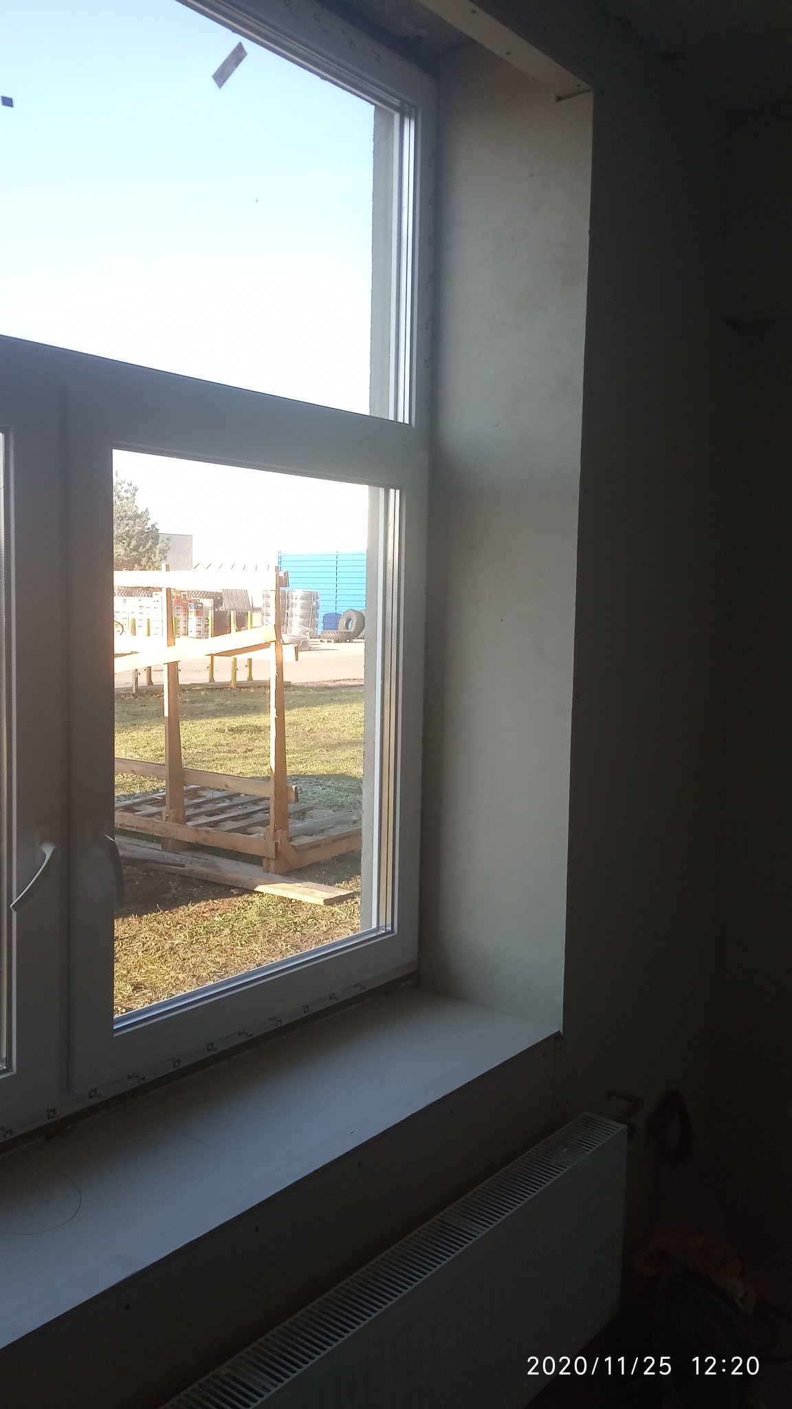 Świeżo osadzony, biały profil okienny w surowym wnętrzu, widok na zewnątrz na palety i niebieski kontener, ściana przygotowana pod wykończenie, widoczny grzejnik.