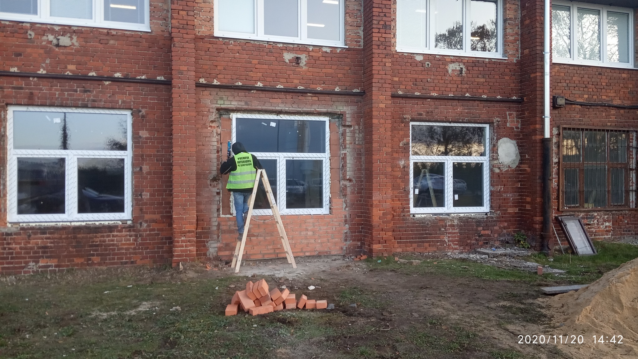 Pracownik w kamizelce, stojący na drabinie, uszczelnia silikonem nowo zamontowane okno w ścianie z czerwonej cegły, obok sterta cegieł i piasku.