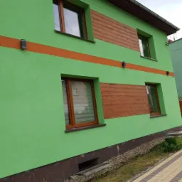 Budynek w Dzierżoniowie z zieloną elewacją, brązowymi oknami i poziomymi panelami imitującymi drewno, oświetlony zewnętrznymi lampami.