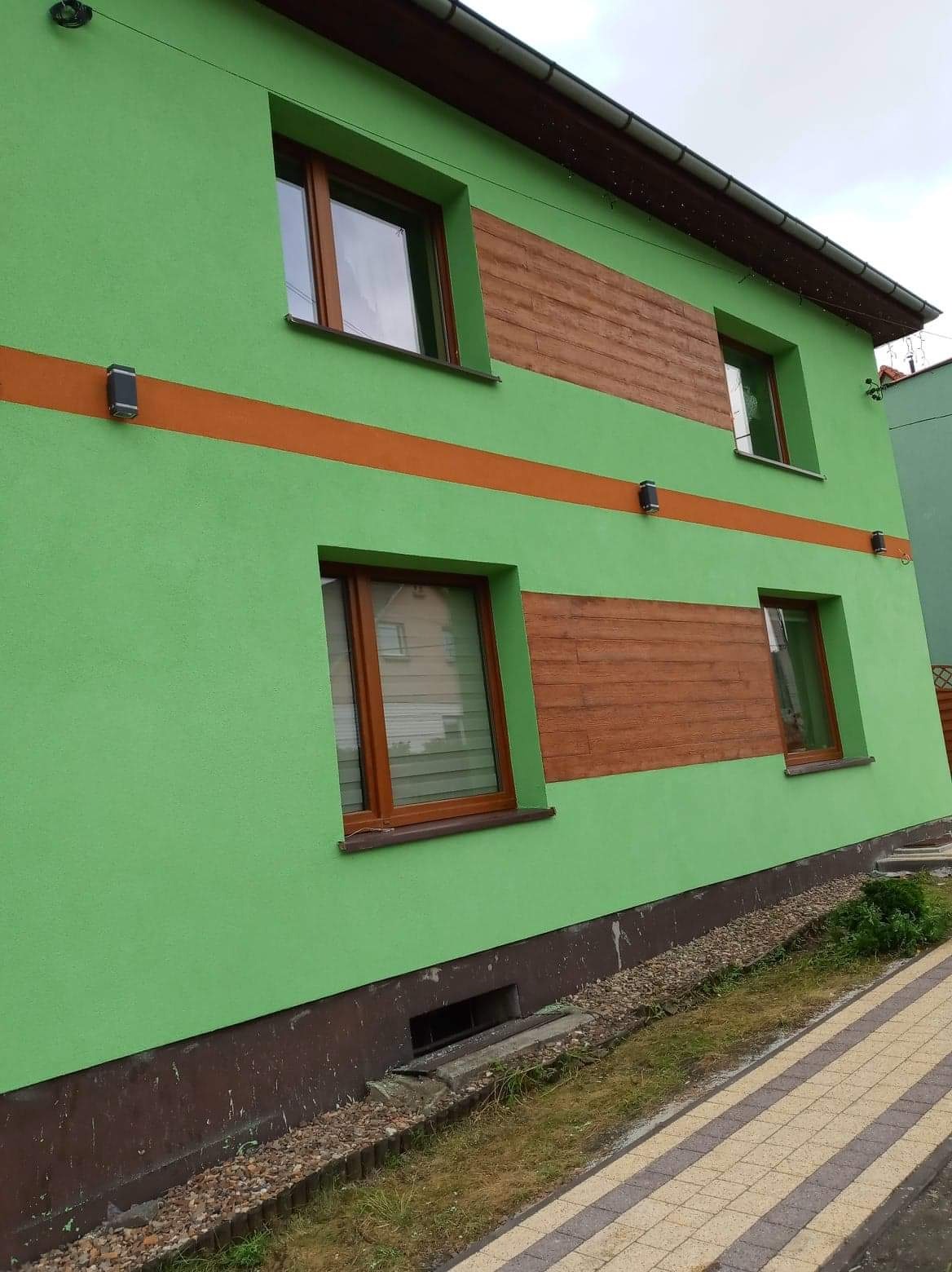 Budynek w Dzierżoniowie z zieloną elewacją, brązowymi oknami i poziomymi panelami imitującymi drewno, oświetlony zewnętrznymi lampami.