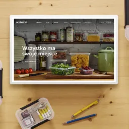 Zdjęcie przedstawia tablet wyświetlający stronę internetową z produktami do przechowywania żywności, otoczony przez pudełka do przechowywania, długopisy i inne przedmioty na drewnianym stole...