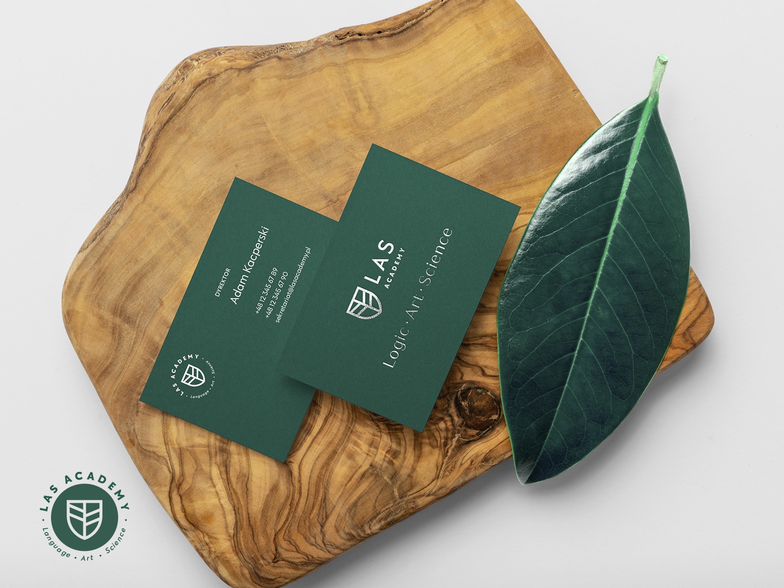 Dwie ciemnozielone wizytówki z białym logo i danymi kontaktowymi na drewnianej desce, obok duży, ciemnozielony liść.