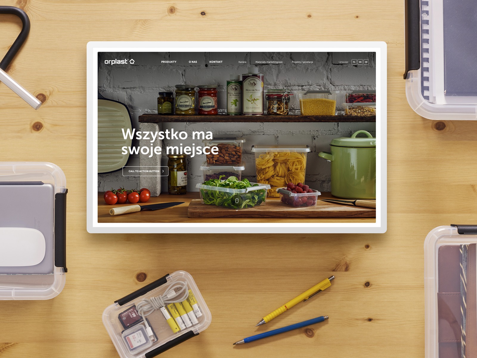 Zdjęcie przedstawia tablet wyświetlający stronę internetową z produktami do przechowywania żywności, otoczony przez pudełka do przechowywania, długopisy i inne przedmioty na drewnianym stole...