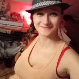 Uśmiechnięta kobieta w kapeluszu, z warkoczem, w beżowej koszulce na tle pokoju z suszarką na pranie i butelką wody.