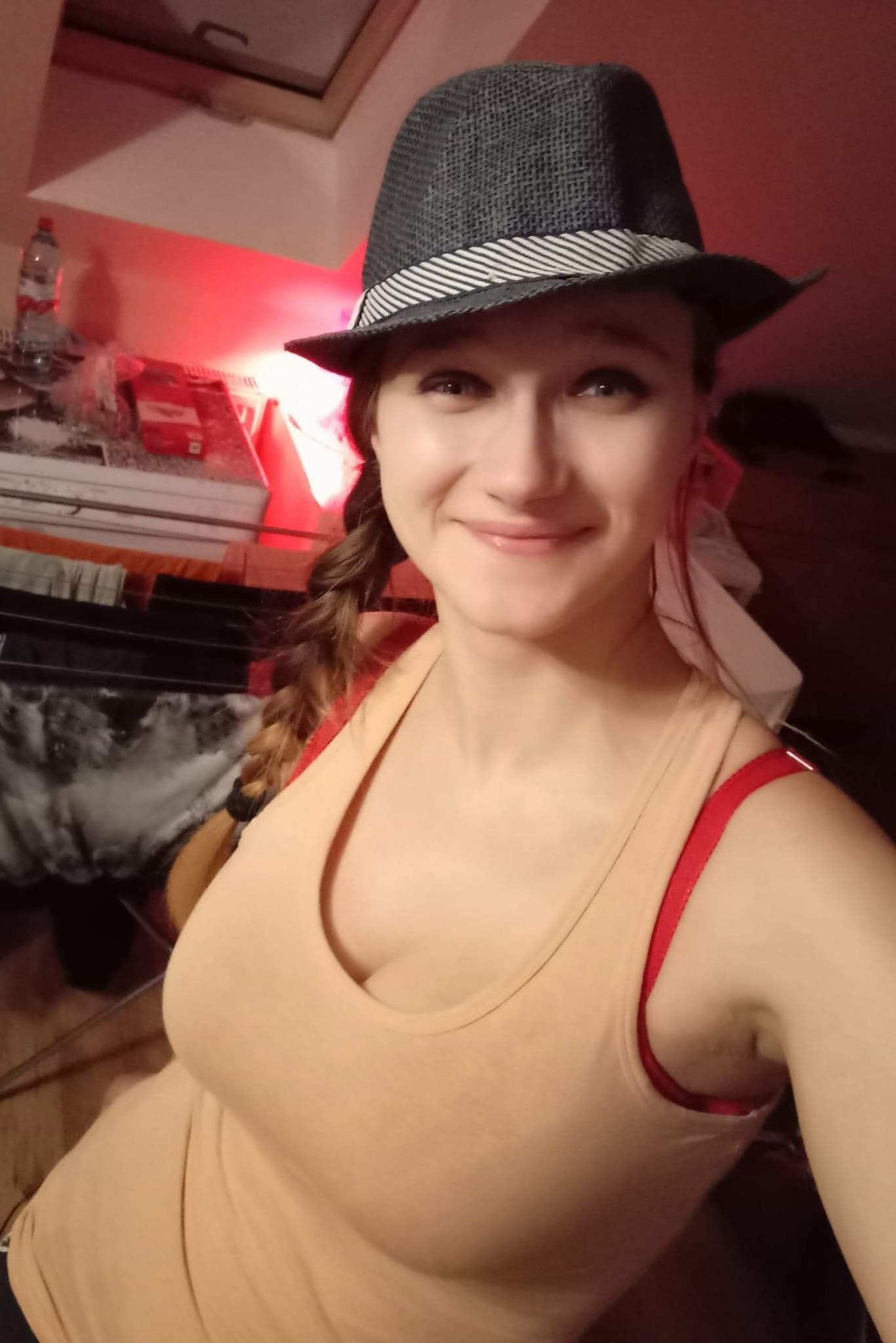 Uśmiechnięta kobieta w kapeluszu, z warkoczem, w beżowej koszulce na tle pokoju z suszarką na pranie i butelką wody.