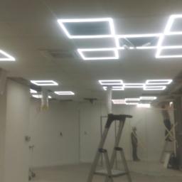 Bart-Tom-Dom - Wnętrze biurowe w trakcie prac wykończeniowych; widoczna drabina, pracownik na rusztowaniu, sufit podwieszany z kwadratowymi lampami LED.