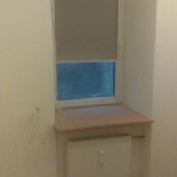 Bart-Tom-Dom - Wnęka okienna z zamontowanym blatem parapetu, roletą w oknie i grzejnikiem pod spodem, ściany w trakcie przygotowania pod malowanie, widoczne gniazdka elektryczne.