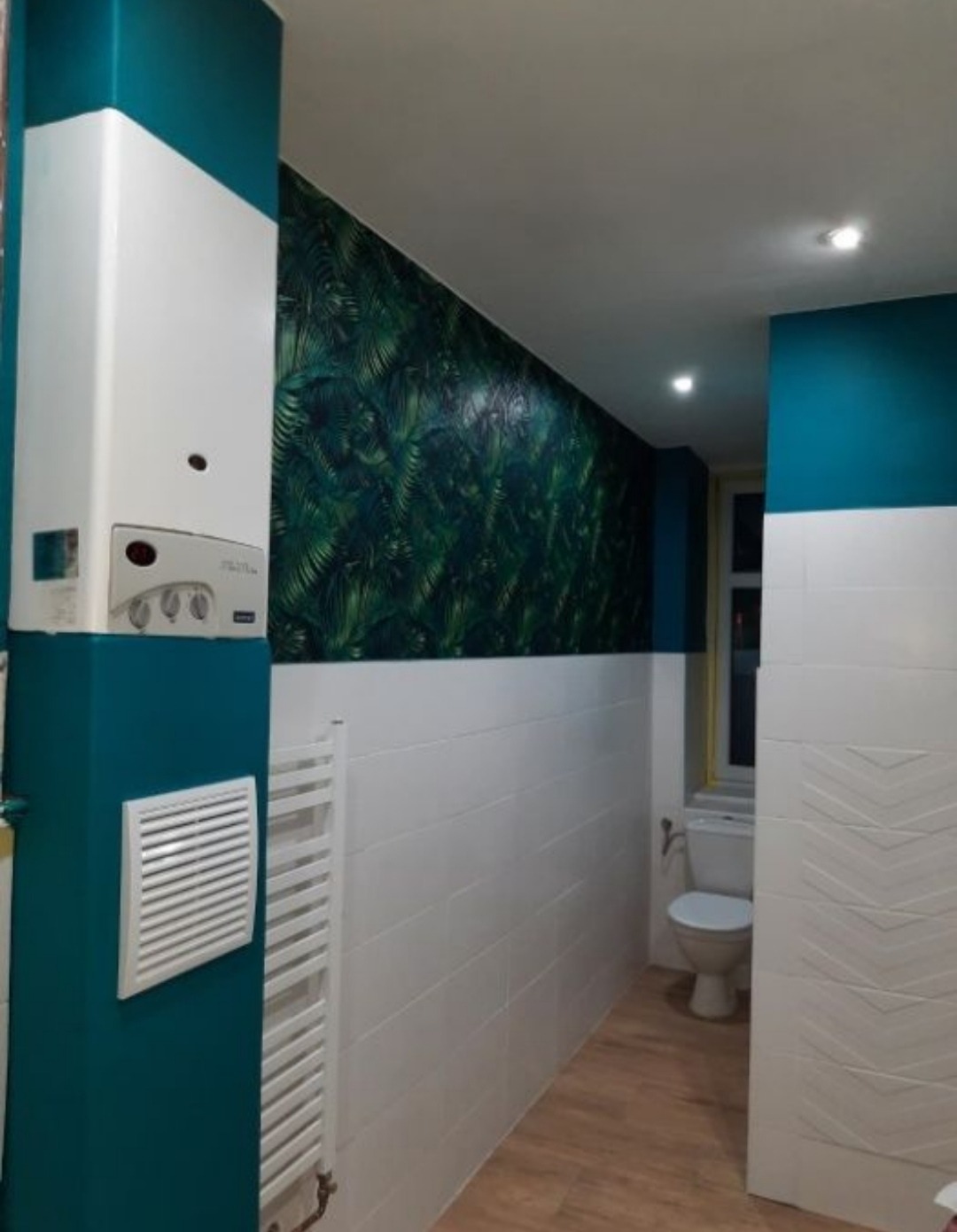 Wykończona łazienka w Poznaniu z białymi płytkami, fragmentem ściany w tropikalne liście i turkusowymi akcentami, widoczny grzejnik drabinkowy i piec gazowy.