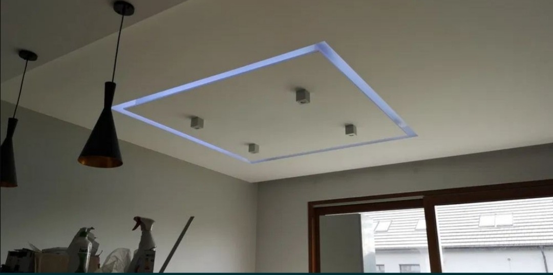 Podwieszany sufit z oświetleniem LED w kształcie kwadratu, uzupełniony trzema punktowymi lampami, widoczne czarne, stożkowe lampy wiszące oraz fragment okna.