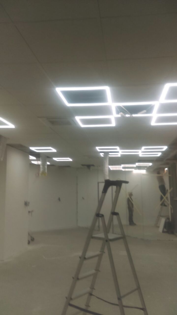 Wnętrze biurowe w trakcie prac wykończeniowych; widoczna drabina, pracownik na rusztowaniu, sufit podwieszany z kwadratowymi lampami LED.