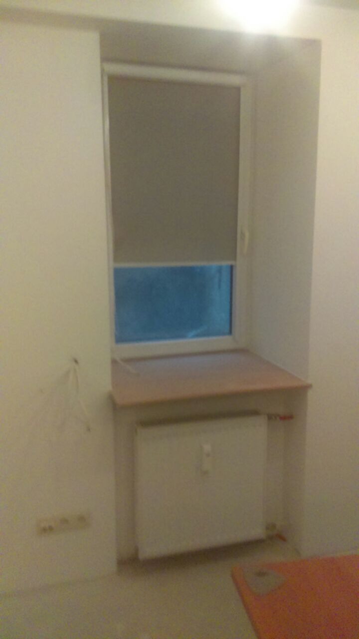 Wnęka okienna z zamontowanym blatem parapetu, roletą w oknie i grzejnikiem pod spodem, ściany w trakcie przygotowania pod malowanie, widoczne gniazdka elektryczne.