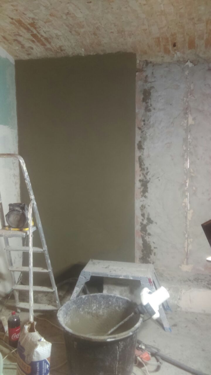 Ściana w trakcie prac wykończeniowych: częściowo wyszpachlowana, z widoczną ceglaną konstrukcją sufitu, drabina, wiadro z zaprawą i narzędzia w pomieszczeniu.