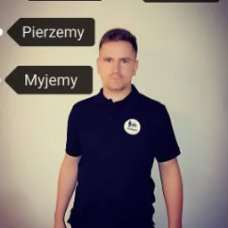Mężczyzna w czarnej koszulce polo z logo firmy ProSteam, na tle ściany z napisem 'Pierzemy' i 'Myjemy' powyżej.