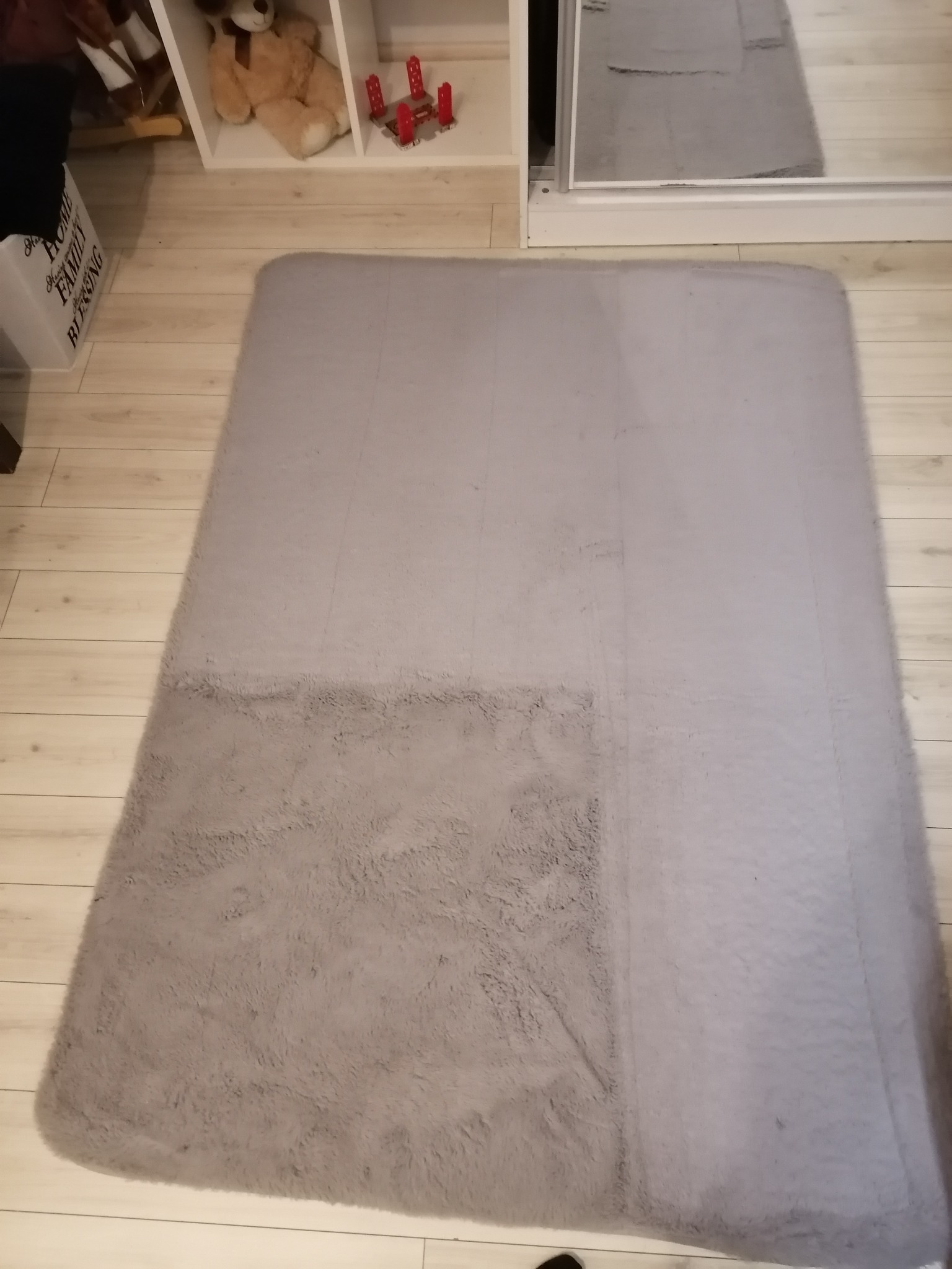 Szary, prostokątny dywan shaggy leżący na panelach podłogowych, widoczne meble w tle.