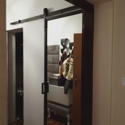 Deco-Stal - Białe przesuwne drzwi w systemie szynowym, odsłaniające fragment przedpokoju z siedziskiem, wieszakiem na ubrania i panelem tapicerowanym.