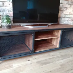 Stylowa szafka RTV z metalową ramą i drewnianymi elementami, z siatkowymi drzwiczkami, na której stoi telewizor, a obok dekoracyjne słoiki z korkami i rośliną, na tle ceglanej ściany.