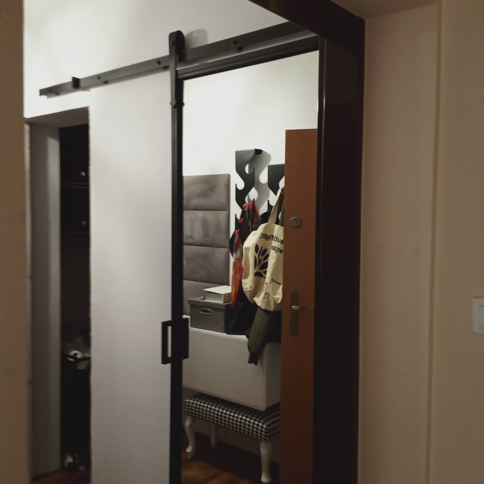 Białe przesuwne drzwi w systemie szynowym, odsłaniające fragment przedpokoju z siedziskiem, wieszakiem na ubrania i panelem tapicerowanym.
