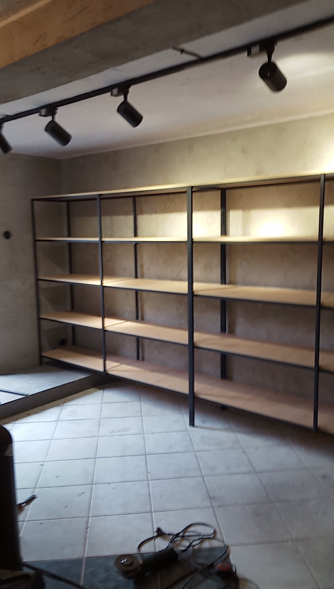 Metalowa konstrukcja regału z drewnianymi półkami w surowym wnętrzu, widoczne narzędzia na podłodze.