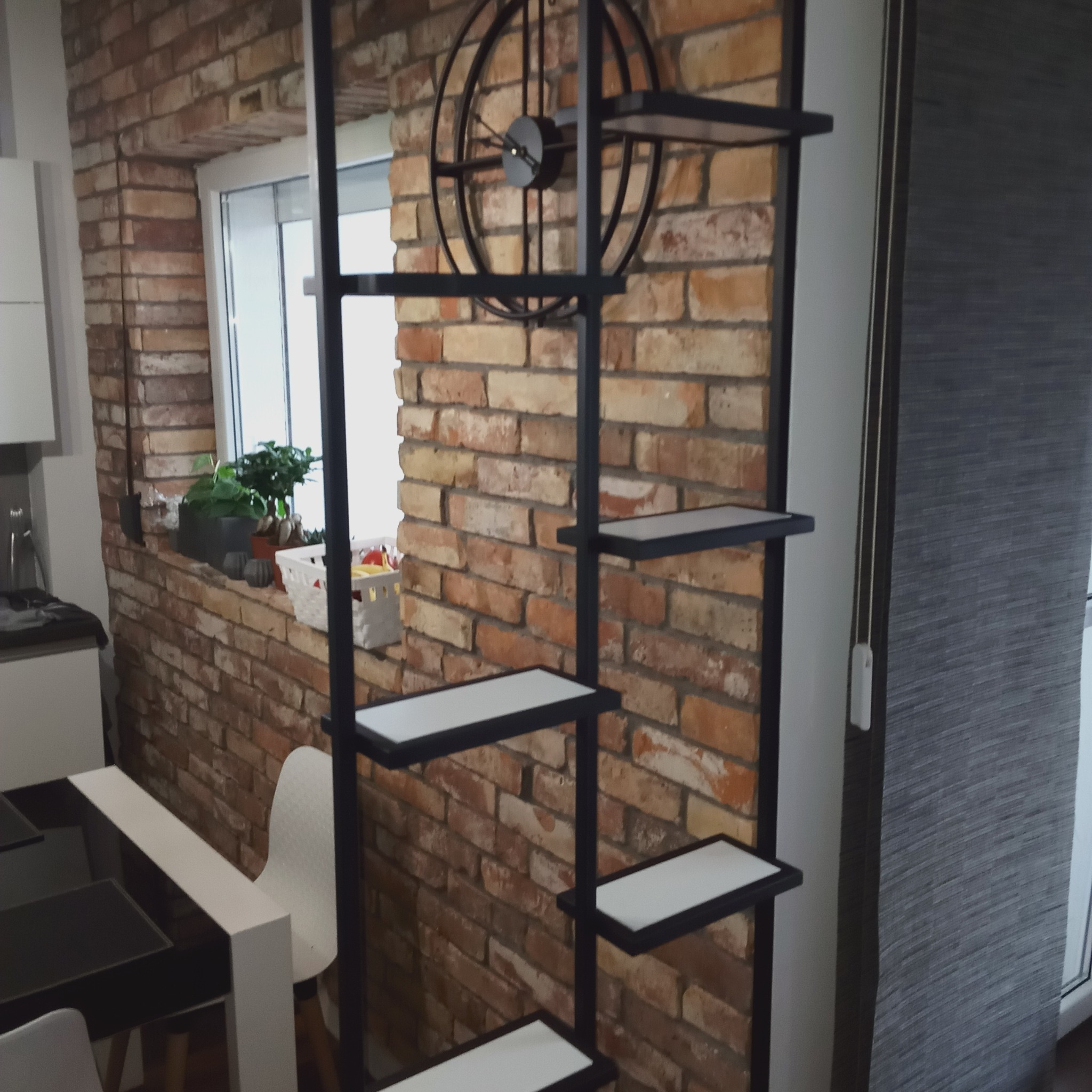 Metalowy regał z półkami na tle ceglanej ściany z zegarem, uzupełnienie aranżacji wnętrza.