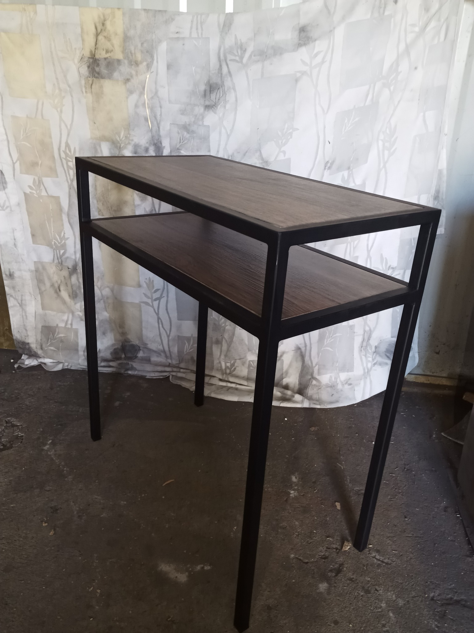 Stolik konsola z dwoma blatami z ciemnego drewna i czarną metalową ramą, na tle wzorzystej tkaniny.