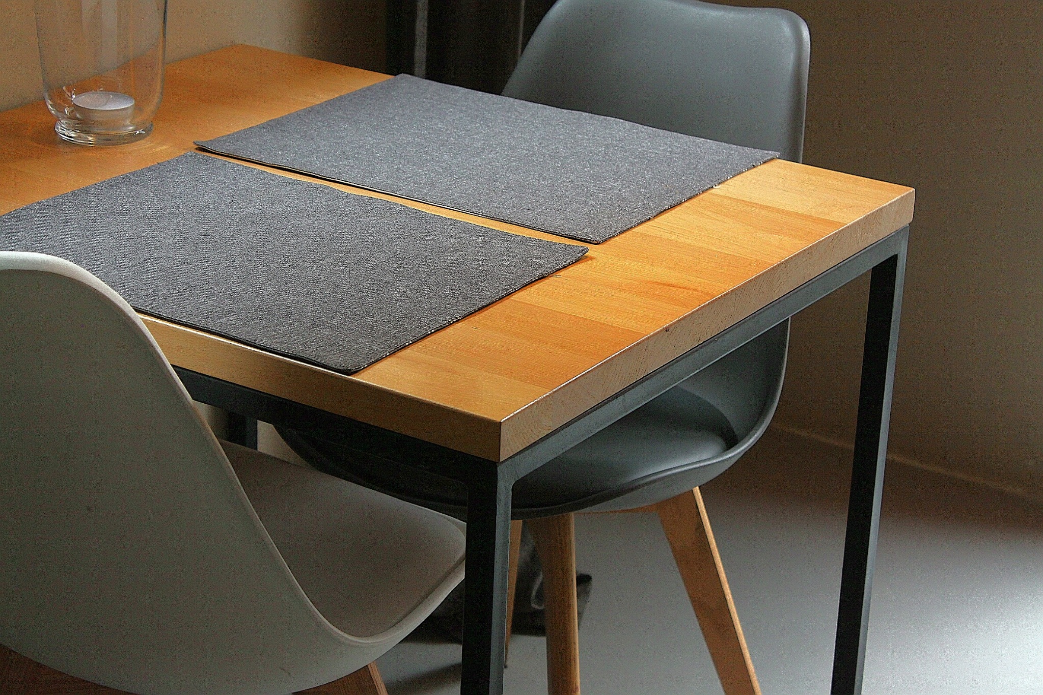 Stół z blatem z jasnego drewna i metalową ramą, nakryty dwoma szarymi podkładkami, otoczony krzesłami w kolorach białym i szarym, z widocznym szklanym naczyniem.