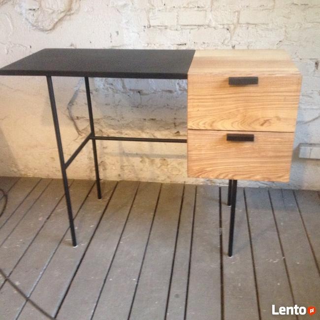 Biurko z blatem z czarnej płyty i drewnianą szafką z dwoma szufladami na metalowych nóżkach, ustawione na szarej podłodze z desek na tle ściany z białej cegły.