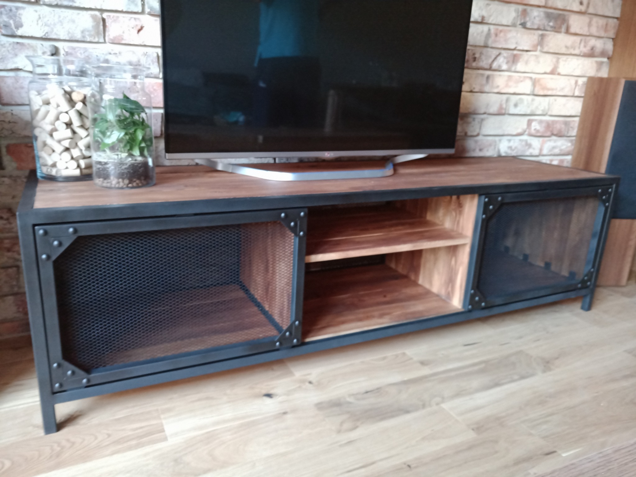 Stylowa szafka RTV z metalową ramą i drewnianymi elementami, z siatkowymi drzwiczkami, na której stoi telewizor, a obok dekoracyjne słoiki z korkami i rośliną, na tle ceglanej ściany.