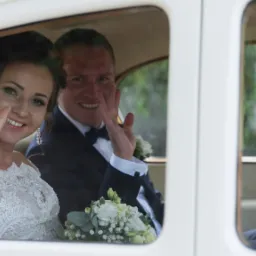 Uśmiechnięta para młoda w klasycznym białym samochodzie, machająca przez okno, bukiet ślubny na kolanach panny młodej, ciemny garnitur pana młodego z muchą.